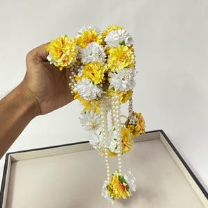 以下が含まれることがあります： 黄色と白の造花とパールの房が付いた花のガーランド。ガーランドはニュートラルな背景に配置され、鮮やかな色と複雑なデザインを際立たせています。お祝いの席に最適です。