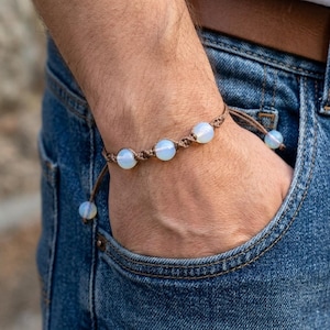 Pode incluir: Uma pulseira de macramé com cinco contas redondas e opalescentes. As contas são enfiadas num cordão castanho, com um fecho de nó deslizante. A pulseira é usada no pulso, com calças de ganga azuis no fundo.