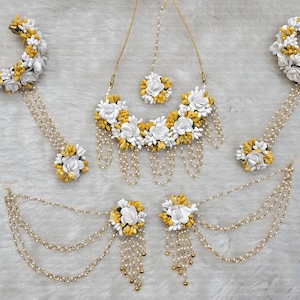 May include: A white and yellow flower jewelry set with a necklace, earrings, bangles, and a maang tikka. The jewelry is made of artificial flowers and beads.
