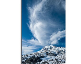 May include: A scenic photograph of a snow-covered mountain under a bright blue sky with wispy white clouds. The mountain's peak is covered in snow, and the lower slopes show dark rock formations. The sky is a vibrant blue, with the clouds adding texture.