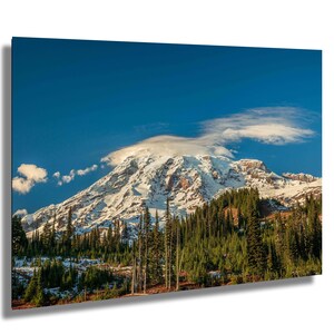 May include: A snow-capped mountain peak with a cloud formation above it. The mountain is surrounded by a forest of evergreen trees.