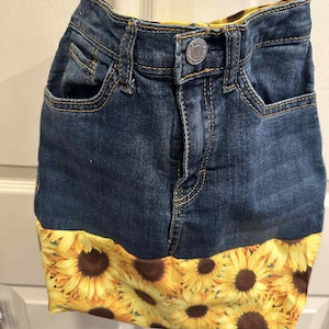 May include: A denim crossbody bag with a sunflower print bottom. The bag is made from repurposed jeans, featuring pockets and a button closure. The bottom of the bag is adorned with a vibrant yellow fabric covered in large sunflowers.
