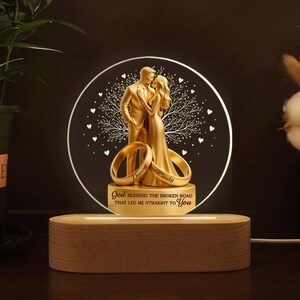May include: A golden-colored wedding night light with a couple embracing, two rings, and the text "God blessed the broken road that led me straight to You." The light sits on a wooden base.