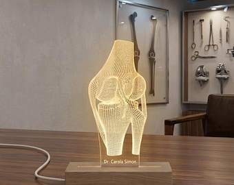 May include: A clear acrylic lamp featuring a glowing, warm-toned wireframe illustration of a human knee joint. The lamp is mounted on a rectangular wooden base with a power cord and includes the engraved text Dr. Carola Simon on the front.