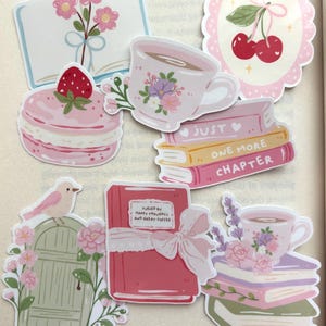 May include: A collection of stickers featuring illustrations of pink flowers in a book, cherries in a scalloped frame, a strawberry macaron, a floral teacup, a stack of books labelled Just One More Chapter, a bird on a door, and a book with a bow.
