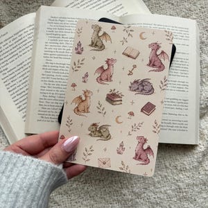 May include: A beige notebook with a repeating pattern of dragons in brown, pink, and grey, alongside books, crystals, and crescent moons. The notebook is held open over an open book.