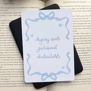 Puede incluir: Un marcapáginas rectangular blanco con un borde decorativo azul claro con lazos en la parte superior e inferior. El texto "crying over fictional characters" está escrito en letra cursiva azul en el centro, acentuado por tres pequeños corazones azules.