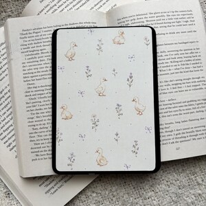 May include: An e-reader cover with a duck and floral pattern in pastel shades. The cover is black and lies on open books. The books are open to pages with text.