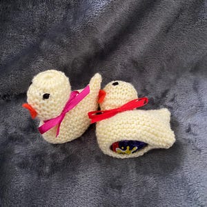 May include: Two knitted yellow duck figurines with pink and red ribbon bows. The ducks are holding chocolate eggs.