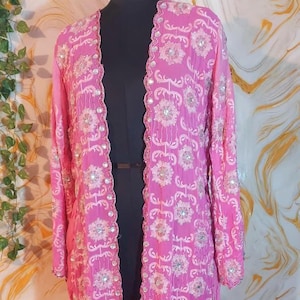 Könnte beinhalten: Eine lange, offen geschnittene, rosa Jacke mit aufwendiger floraler Stickerei und funkelnden Verzierungen. Die Jacke wird auf einer schwarzen Schaufensterpuppe präsentiert, die ihr fließendes Design und ihre detaillierte Verarbeitung hervorhebt. Die Ärmel sind lang und die Kanten sind gewellt.