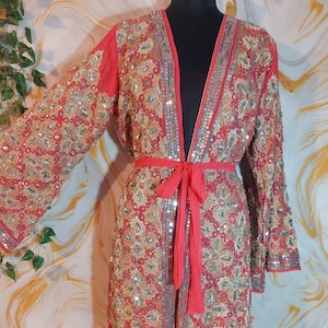 Könnte beinhalten: Langer, offen geschnittener Robe mit aufwendiger Gold- und Silberstickerei auf rotem Hintergrund. Die Robe hat einen passenden roten Bindegürtel und wird auf einer schwarzen Schaufensterpuppe präsentiert. Die Ärmel sind lang und weit, mit Paillettenbesatz.