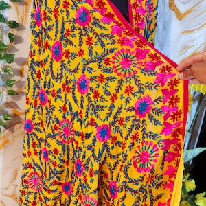 May include: A bright yellow textile with detailed embroidery. The design includes pink and blue floral patterns with red accents. The edges are trimmed with a red border. The textile is likely a scarf or shawl.