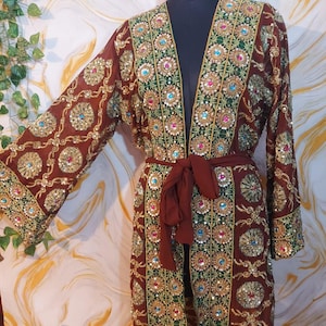 Könnte beinhalten: Ein brauner, offen geschnittener Robe mit aufwendiger Blumenstickerei und Paillettenverzierungen. Die Robe hat lange Ärmel und einen passenden Bindegürtel. Die Stickerei umfasst goldene, grüne, blaue und rosa Akzente und schafft ein dekoratives und elegantes Design.