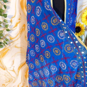 May include: A bright blue shawl with detailed embroidery. The design includes circular and cross-shaped patterns in red, yellow, and white. The shawl has a border with small reflective embellishments and a yellow trim.