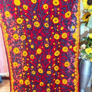 May include: A red textile with an embroidered floral pattern. The design features yellow flowers, blue stems, and paisley shapes. The edges are trimmed with a yellow border, creating a striking visual contrast.
