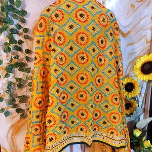 May include: A yellow shawl with a geometric pattern of diamonds and circles in red, blue, and black. The shawl has a black border with mirrored accents and is displayed on a metal stand. The background includes greenery and sunflowers.
