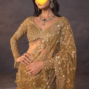 May include: A gold-coloured saree featuring intricate embroidery with sequins and beads. The long-sleeved blouse has a V-neckline and matching embellishments. A necklace with green and clear stones and matching earrings are worn with the garment.