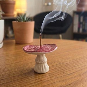 Könnte beinhalten: Ein Räucherstäbchenhalter aus Keramik in Form eines Pilzes mit rotem Hut, weißen Punkten und beigem Stiel. Ein dünnes Räucherstäbchen steckt in der Mitte des Pilzhutes und gibt eine sichtbare weiße Rauchfahne ab, die nach oben steigt.