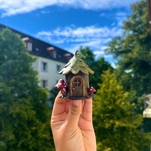 May include: A miniature fairy house with a brown door, green roof, and two red mushroom accents. The whimsical house is held up against a backdrop of green trees and a blue sky.