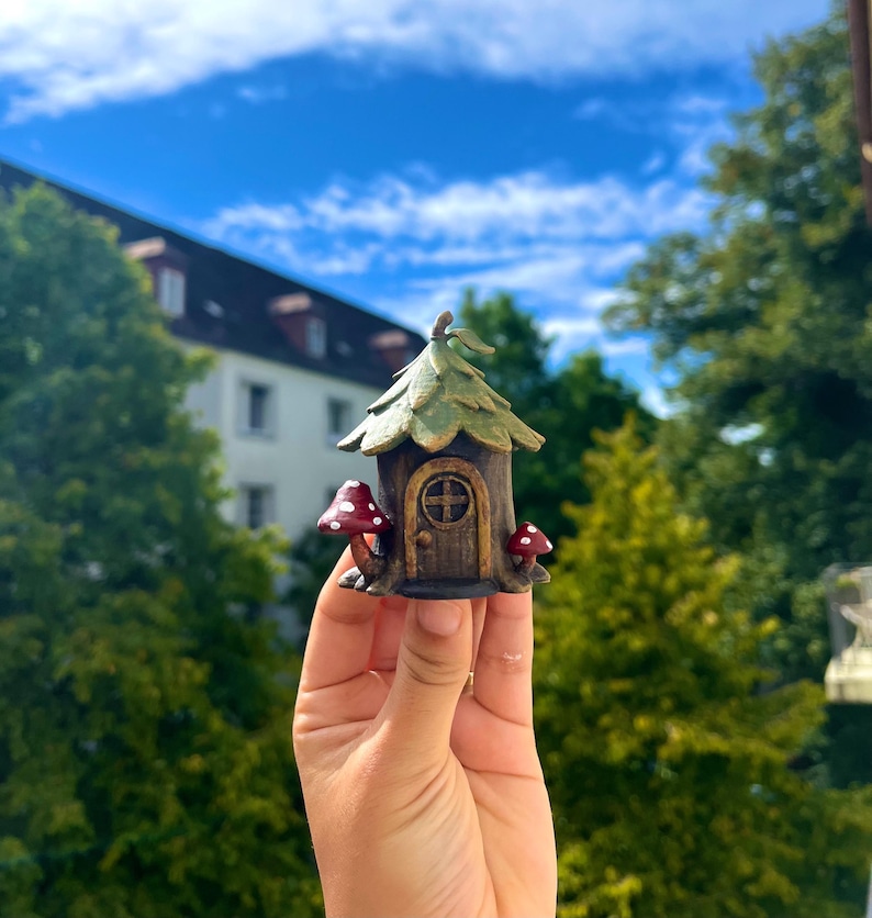 May include: A miniature fairy house with a brown door, green roof, and two red mushroom accents. The whimsical house is held up against a backdrop of green trees and a blue sky.