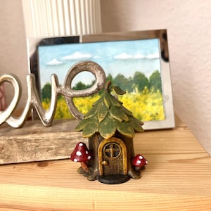May include: A whimsical fairy house figurine with a green leaf-like roof, a brown door, and two red and white mushroom accents. The miniature house is set on a wooden surface, with a framed landscape painting and a metal "LOVE" sign in the background.