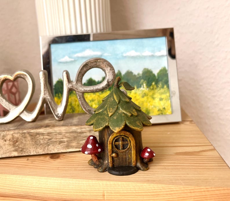 May include: A whimsical fairy house figurine with a green leaf-like roof, a brown door, and two red and white mushroom accents. The miniature house is set on a wooden surface, with a framed landscape painting and a metal "LOVE" sign in the background.