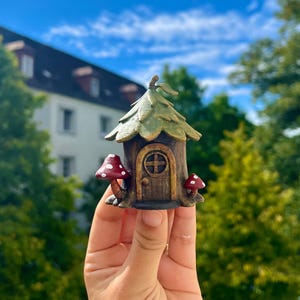 May include: A miniature fairy house with a brown wooden door and a green leaf-shaped roof. Two red mushrooms with white spots flank the entrance. The house is held against a backdrop of trees and a blue sky.