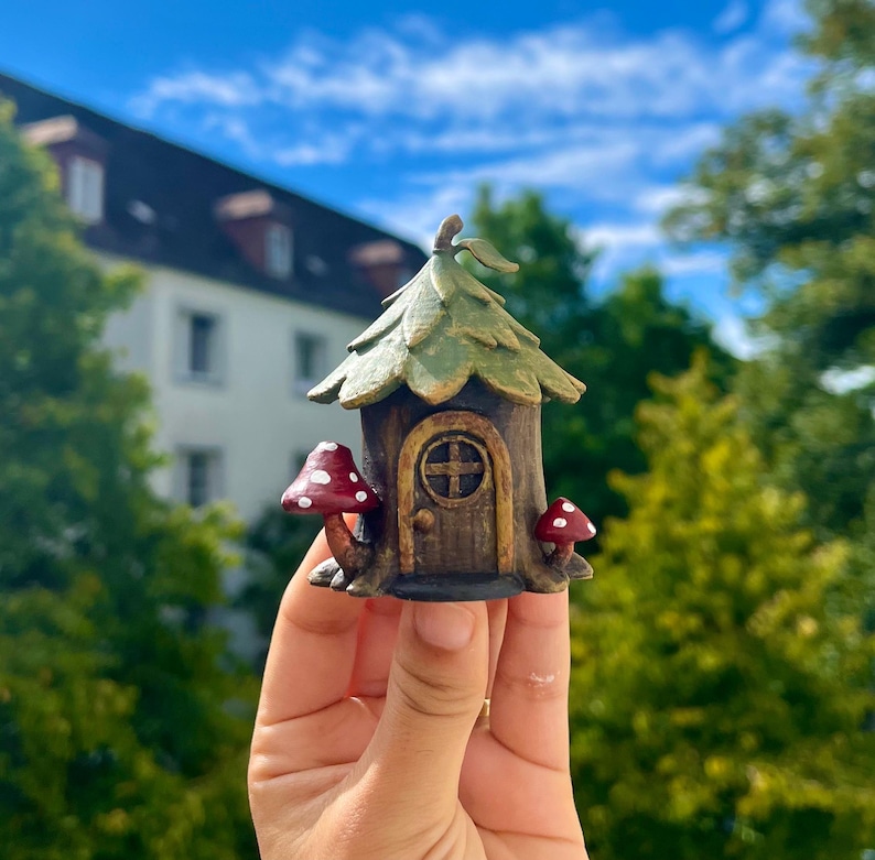 May include: A miniature fairy house with a brown wooden door and a green leaf-shaped roof. Two red mushrooms with white spots flank the entrance. The house is held against a backdrop of trees and a blue sky.