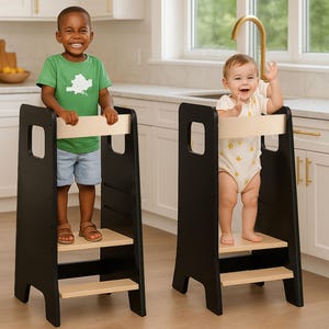 Può includere: Due sgabelli neri e in legno naturale con bambini in piedi sopra. Gli sgabelli hanno una piattaforma e una barra di sicurezza. Un bambino indossa una maglietta verde e pantaloncini blu, l'altro un body bianco con stelle gialle.
