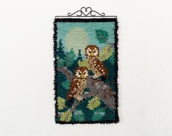 Op de afbeelding: Een vintage wandtapijt met twee bruine uilen die op een tak zitten in een bosrijke omgeving. Het tapijt heeft een blauwe, groene en bruine kleurstelling en is omlijst met een zwarte rand.