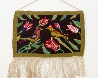 Op de afbeelding: Een geweven wandkleed met een donkerbruine achtergrond met twee gele vogels en roze bloemen met groene bladeren. Het kleed heeft een groene rand en een franje van natuurlijke vezels.