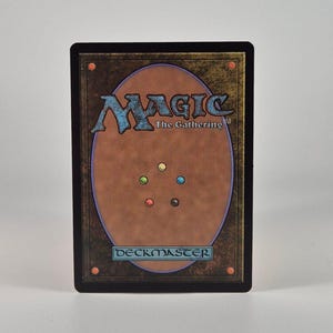 May include: A rectangular Magic The Gathering trading card featuring a brown background with a large central oval. The card displays the text Magic The Gathering and DECKMASTER, along with five small colored circles in yellow, blue, red, black, and green.