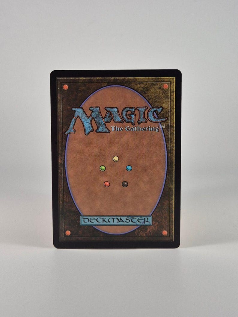 May include: A rectangular Magic The Gathering trading card featuring a brown background with a large central oval. The card displays the text Magic The Gathering and DECKMASTER, along with five small colored circles in yellow, blue, red, black, and green.