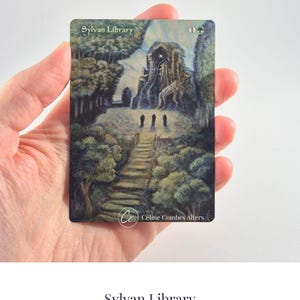 May include: A rectangular trading card titled Sylvan Library featuring a painted illustration of a stone structure, trees, and three dark figures on a path. The card includes the text Sylvan Library and Céline Combes Alters at the bottom.