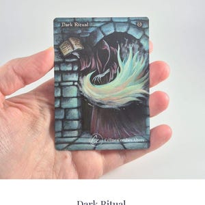 Può includere: Una carta collezionabile rettangolare intitolata Dark Ritual che raffigura una figura incappucciata con una tunica scura sotto un arco di pietra. La figura tiene un libro aperto ed emette un'energia vorticosa di colore verde pallido e giallo. Il testo Céline Combes Alters è stampato in basso.