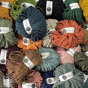 May include: A collection of multiple skeins of Bobbiny cotton cord in various colours including mustard yellow, olive green, dark blue, black, terracotta, beige, dusty pink, and brown. Each skein features a white paper label with the brand name.