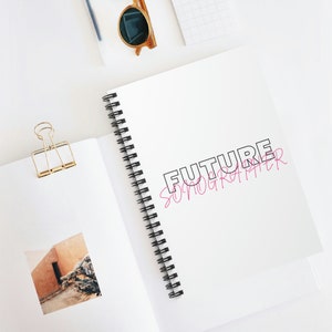 Puede incluir: Una libreta blanca de espiral con el texto "FUTURE SONOGRAPHER" escrito en negro y rosa en la portada.