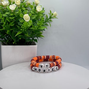 May include: Two bracelets with orange and maroon beads, stacked on top of each other. The bottom bracelet has white beads with the letters "VT" and "HOKIES" spelled out.