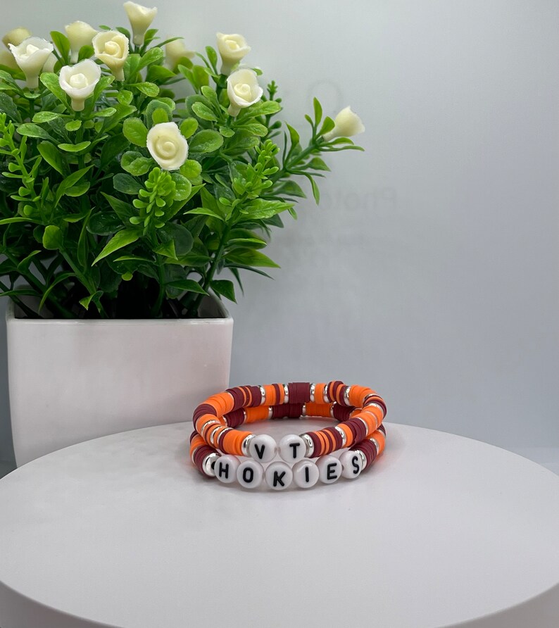 May include: Two bracelets with orange and maroon beads, stacked on top of each other. The bottom bracelet has white beads with the letters "VT" and "HOKIES" spelled out.