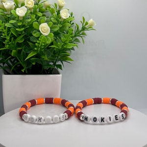May include: Two beaded bracelets with the word "HOKIES" spelled out in white beads. The bracelets are made with maroon, orange, and white beads.