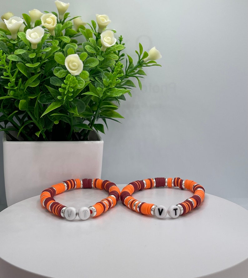 May include: Two orange, maroon, and silver beaded bracelets with white letter beads spelling "VT".