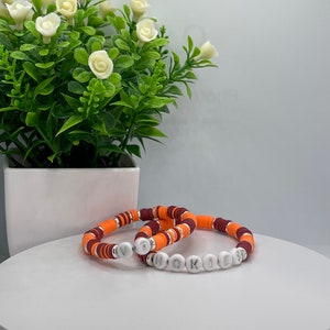 May include: Two bracelets with orange, maroon, and white beads. The bracelets spell out "VT" and "HOKIES".