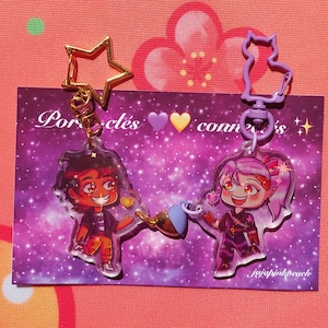 Puede incluir: Dos encantos de personajes en llaveros con un fondo de galaxia púrpura. Los encantos son de dos personajes de dibujos animados, uno con cabello castaño y una camisa azul y el otro con cabello rosado y una camisa morada. El texto en la tarjeta dice "Los portales nos conectan".