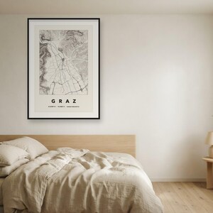 May include: A framed wall art print featuring a detailed black and white topographic map of Graz, Austria. The print includes the city name GRAZ in bold capital letters at the bottom, along with geographic coordinates and elevation data.