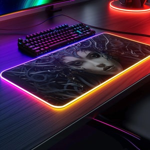 Könnte beinhalten: Rechteckiges Gaming-Mauspad mit einem Frauengesicht-Design und einem leuchtenden LED-Rand. Die Tastatur ist schwarz mit mehrfarbigen Lichtern. Das Mauspad liegt auf einem dunklen Holzschreibtisch.