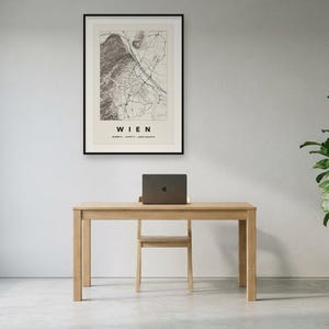 May include: A framed wall art print featuring a detailed black and white map of Vienna, Austria, with the text WIEN and geographic coordinates printed below the map. The print is displayed in a thin black frame on a plain light-coloured wall.