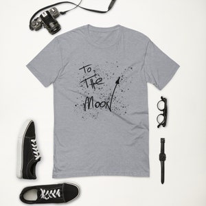 May include: A heather grey t-shirt with a black graphic print that says "To the Moon" with a black arrow pointing upwards. The graphic is surrounded by black splatters.