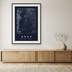 May include: A framed wall art print featuring a dark blue map of Graz, Austria. The map displays white street lines and topographic contours. Below the map, the text reads GRAZ, followed by the coordinates 47.0707 N, 15.4395 E.