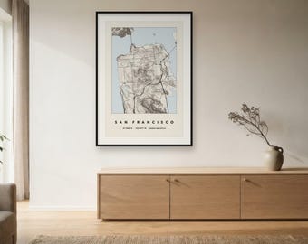 May include: A framed wall art print featuring a topographic map of San Francisco. The map displays detailed terrain lines in shades of brown against a light blue water background. The text reads SAN FRANCISCO with coordinates 37.7749° N, 122.4194° W.