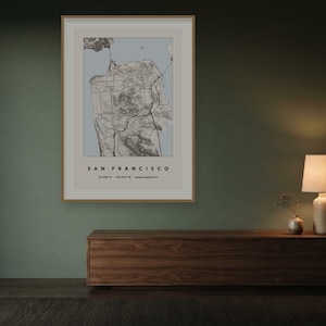 May include: A framed wall art print featuring a topographic map of San Francisco. The map displays detailed terrain lines in black against a light blue background, with the text SAN FRANCISCO and coordinates printed in black at the bottom.
